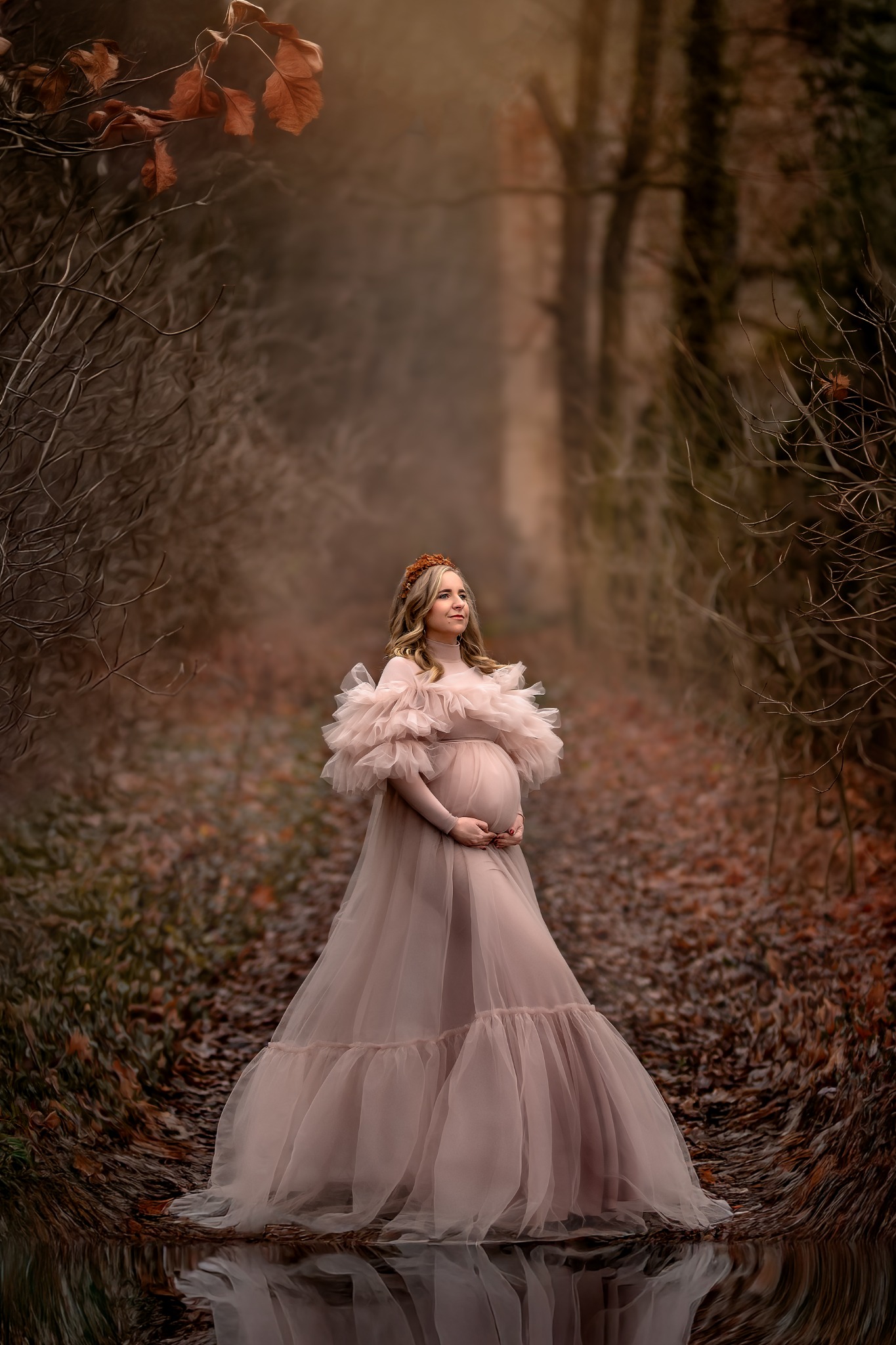 Kobieta w ciąży, w długiej, pudrowo-różowej sukni z falbanami, pozuje w lesie na tle ścieżki z opadłych liści, z koroną z brązowych kwiatów na głowie.
