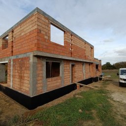 Perfect House - Wznoszony dwukondygnacyjny budynek z czerwonej cegły ceramicznej Porotherm, z widoczną hydroizolacją fundamentów i oknami bez stolarki, na tle pochmurnego nieba i zaparkowanego białego busa z logo...