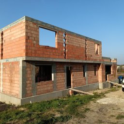 Perfect House - Surowy stan dwukondygnacyjnego domu murowanego z czerwonej cegły Porotherm z widocznymi otworami okiennymi i drzwiowymi, na betonowym fundamencie, z zaparkowanym białym busem firmy budowlanej...