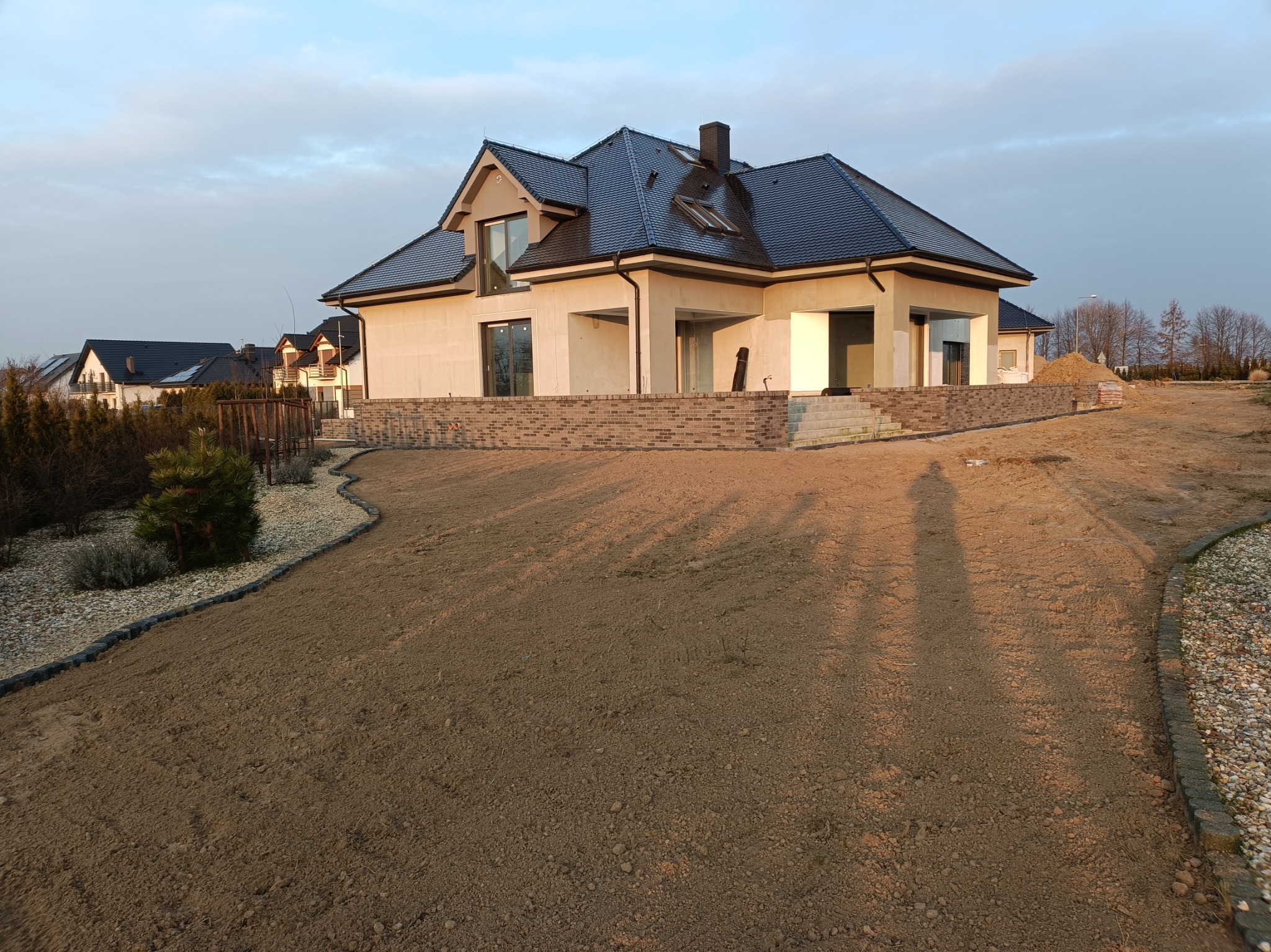 Niedokończony dom jednorodzinny z ciemną dachówką i wstawionymi oknami, otoczony wyrównanym terenem i elementami małej architektury ogrodowej, w tle inne domy.