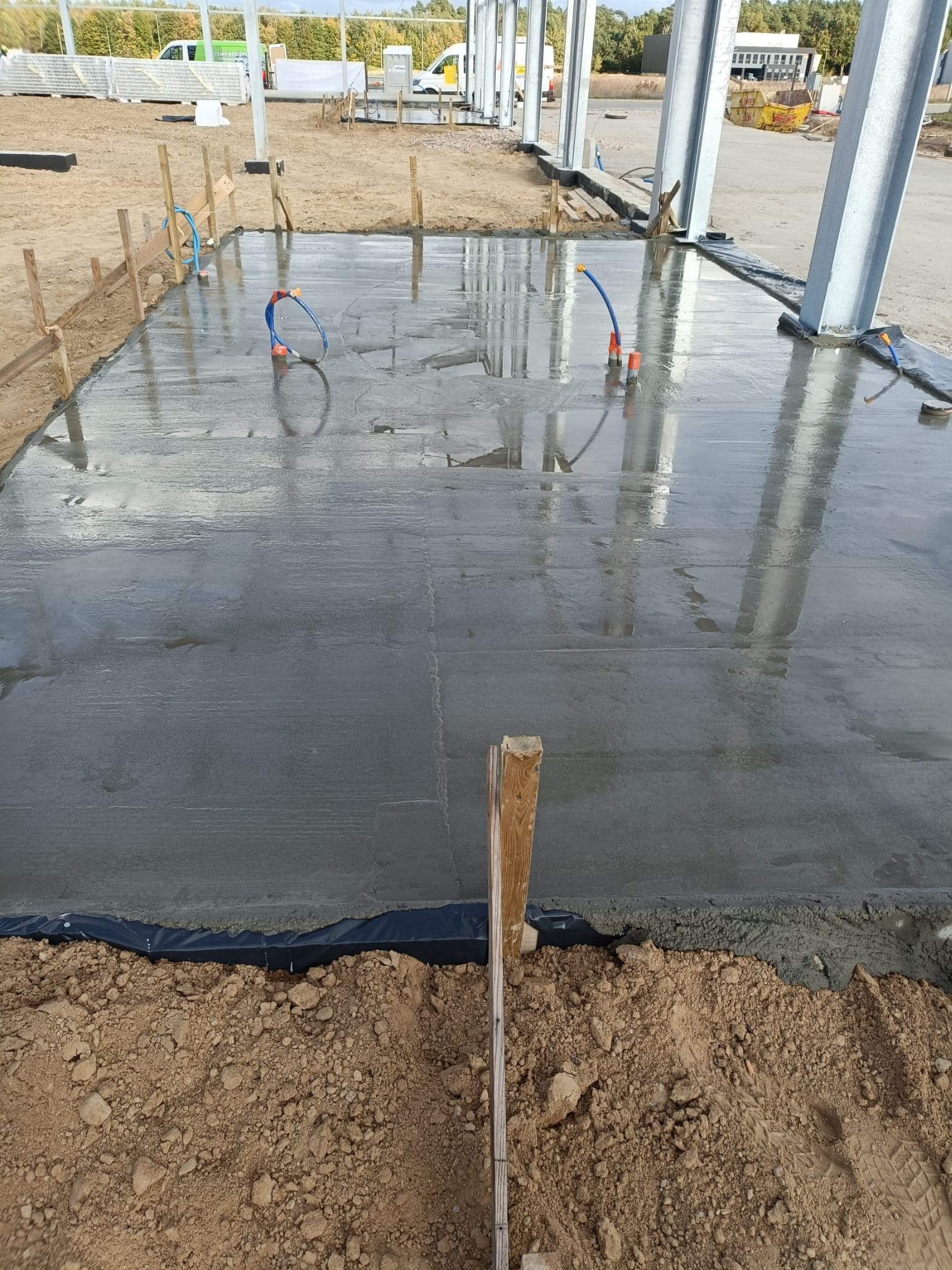 Wylany, mokry beton na fundamencie z widocznymi rurami instalacyjnymi i drewnianym szalunkiem, przygotowany pod budowę hali stalowej.