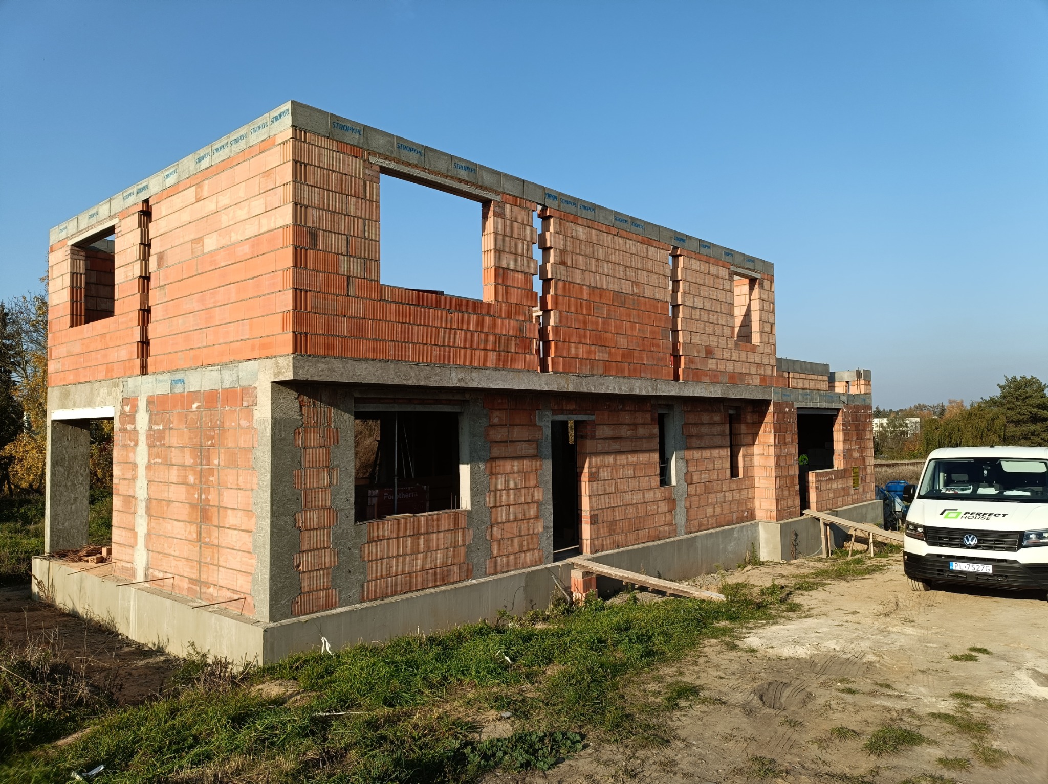 Surowy stan dwukondygnacyjnego domu murowanego z czerwonej cegły Porotherm z widocznymi otworami okiennymi i drzwiowymi, na betonowym fundamencie, z zaparkowanym białym busem firmy budowlanej...