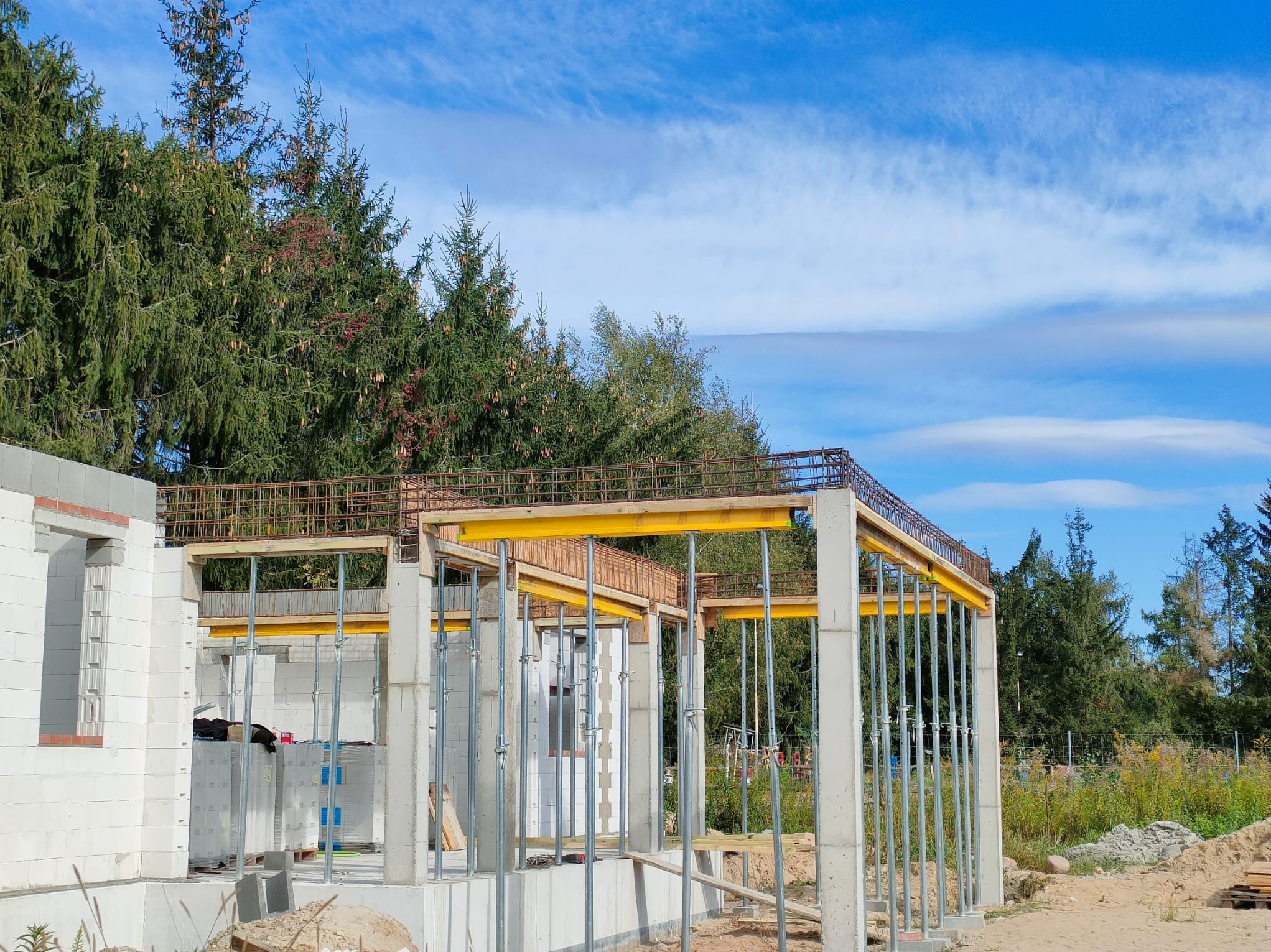 Wznoszenie szkieletu domu z betonu komórkowego, widoczne podpory szalunkowe i zbrojenie stropu na tle lasu i błękitnego nieba.