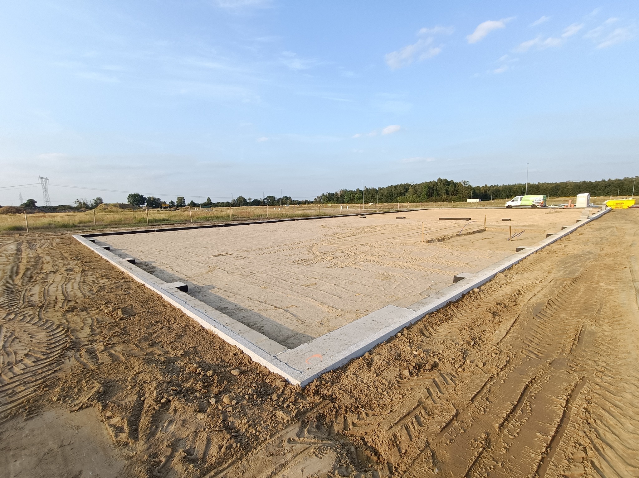Wylany fundament pod budowę domu jednorodzinnego, widoczne ślady opon na piasku, w tle drzewa i błękitne niebo.