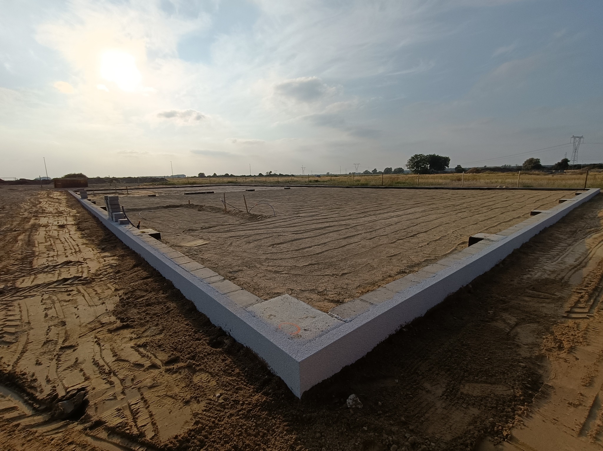 Fundamenty domu jednorodzinnego w trakcie budowy, z widocznym zarysem ścian i podsypką piaskową, na tle zachodzącego słońca i pól.