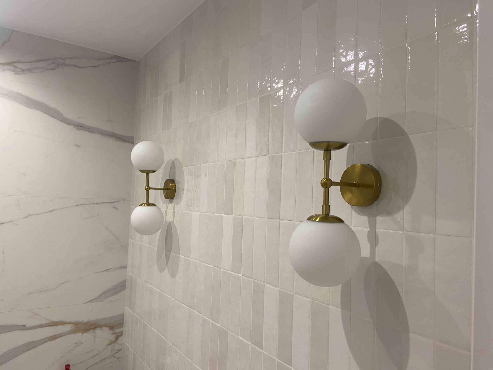 Dwie nowoczesne lampy ścienne z białymi kulistymi kloszami i złotymi mocowaniami na ścianie wyłożonej białymi, błyszczącymi, pionowo ułożonymi płytkami. Po lewej stronie ściana z marmurowym wzorem.