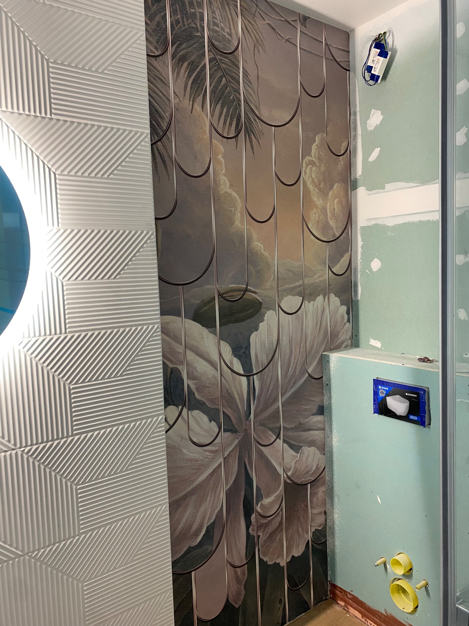 Ściana łazienki z fragmentem lustra, dekoracyjną białą płytką 3D i tapetą z motywem kwiatowym, widoczne elementy instalacji sanitarnej w trakcie montażu.