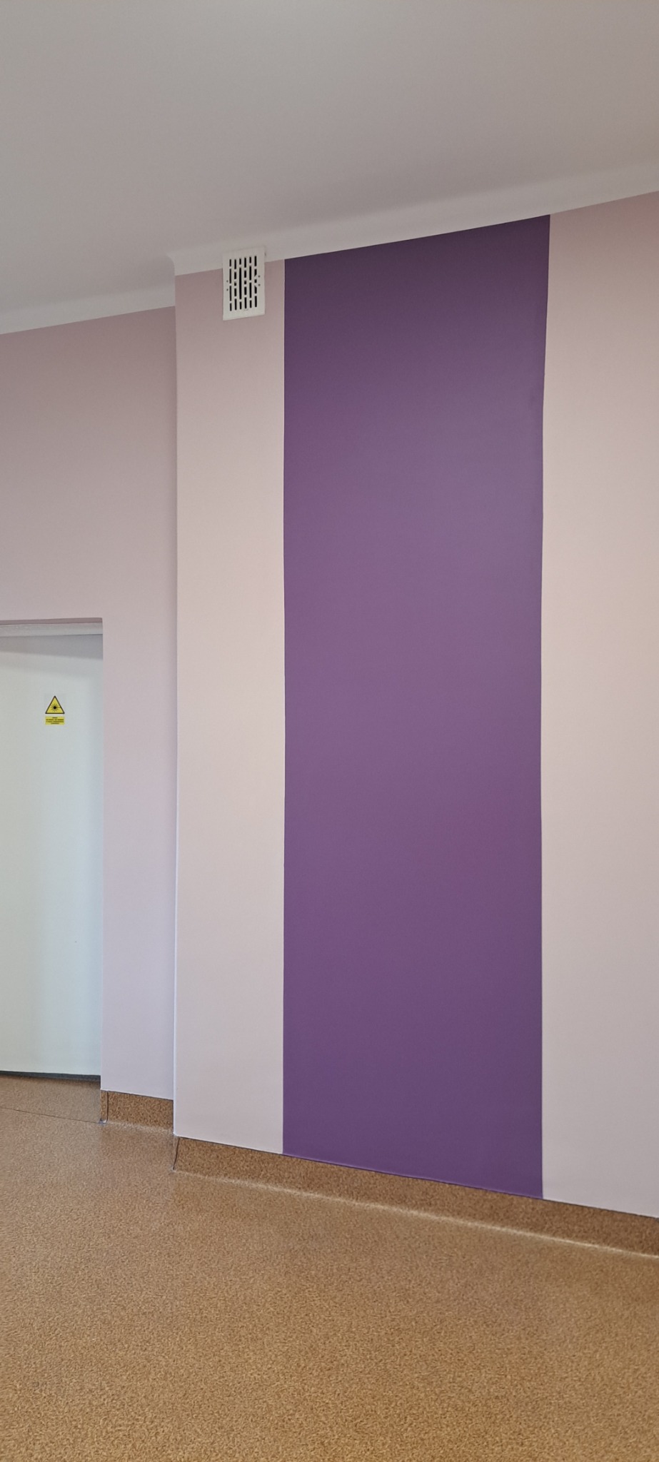 Ściana pomalowana w pasy w odcieniach fioletu i jasnego różu, widoczna kratka wentylacyjna i drzwi z symbolem ostrzegawczym.