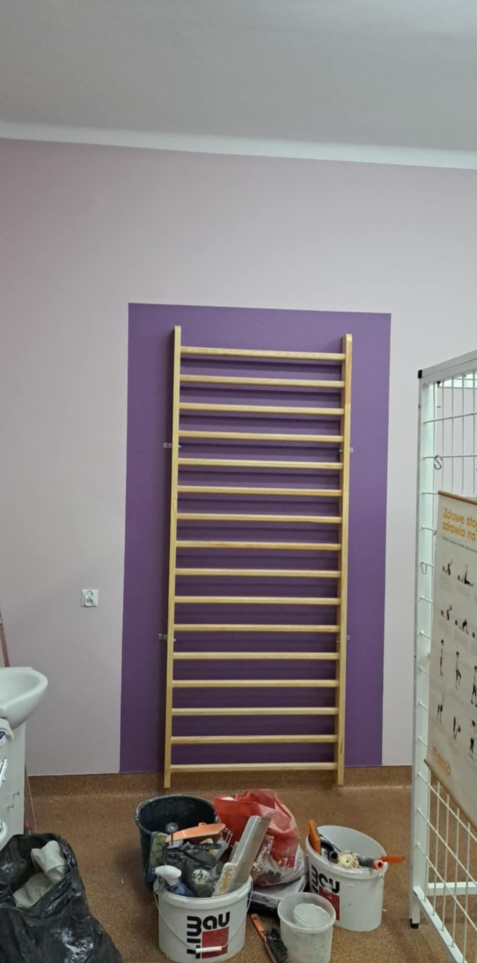 Ściana pomalowana na fioletowo z drabinką gimnastyczną i narzędziami malarskimi, widoczne wiadra z farbą i szpachelki.