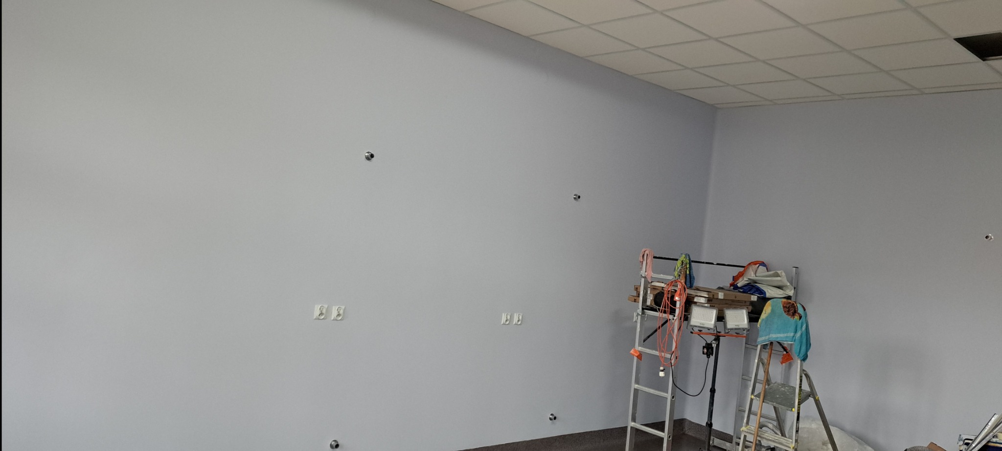Świeżo pomalowane biuro z widocznymi puszkami instalacyjnymi i gniazdkami, przygotowane do dalszych prac wykończeniowych; drabiny i sprzęt malarski w rogu.