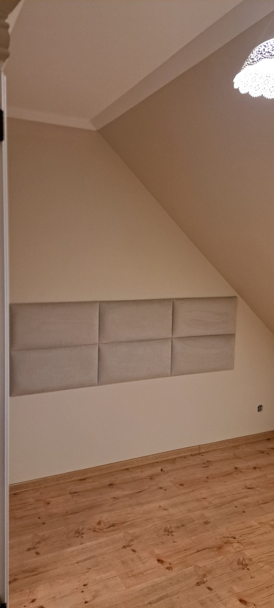 Sześć beżowych paneli tapicerowanych na ścianie ze skosem, drewniana podłoga, widok z wnętrza przez otwarte drzwi.