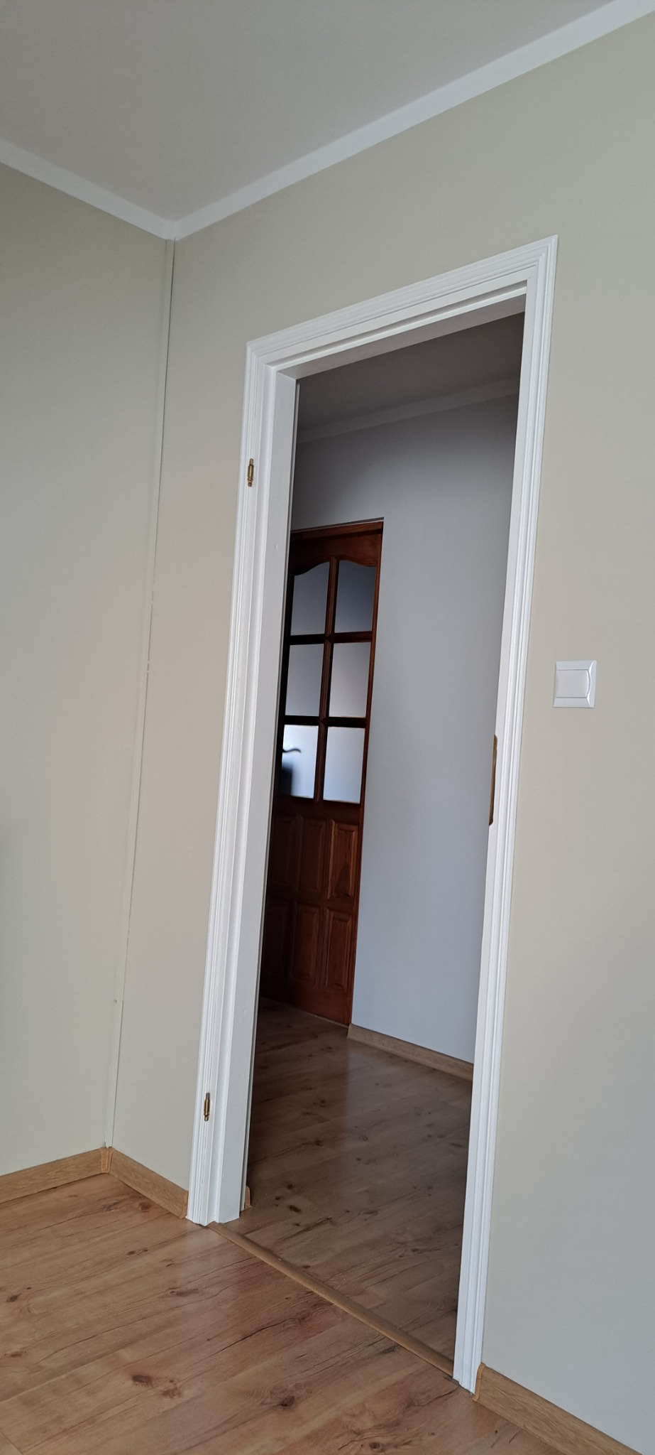 Wnętrze mieszkania po remoncie: biała ościeżnica drzwiowa, świeżo pomalowane ściany w odcieniach beżu, widoczna podłoga laminowana i drewniane drzwi pokojowe z szybami w głębi.