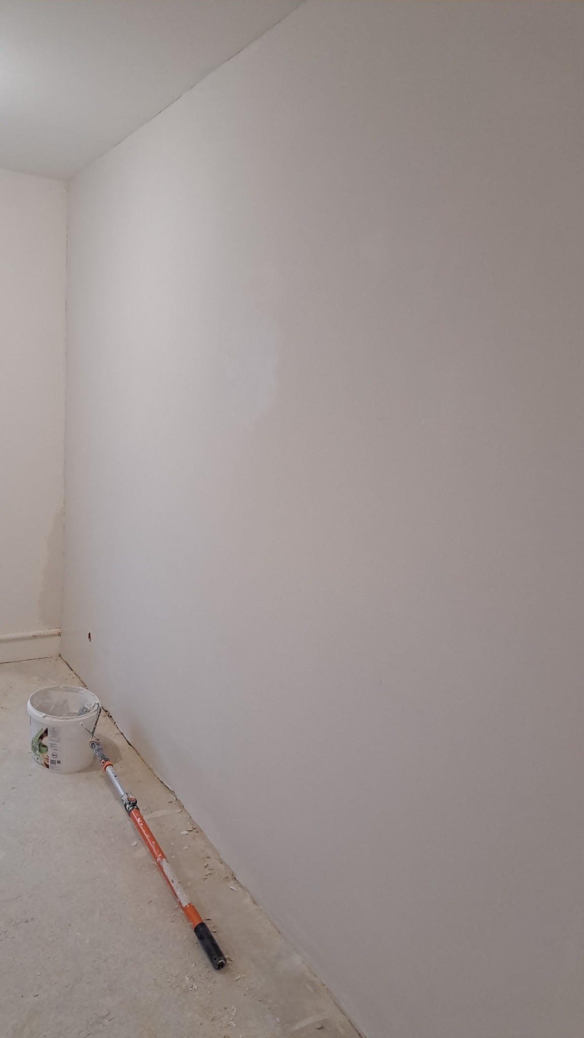 Świeżo pomalowana, jasna ściana w rogu pokoju z widocznym wałkiem malarskim i wiadrem z farbą przy podłodze.