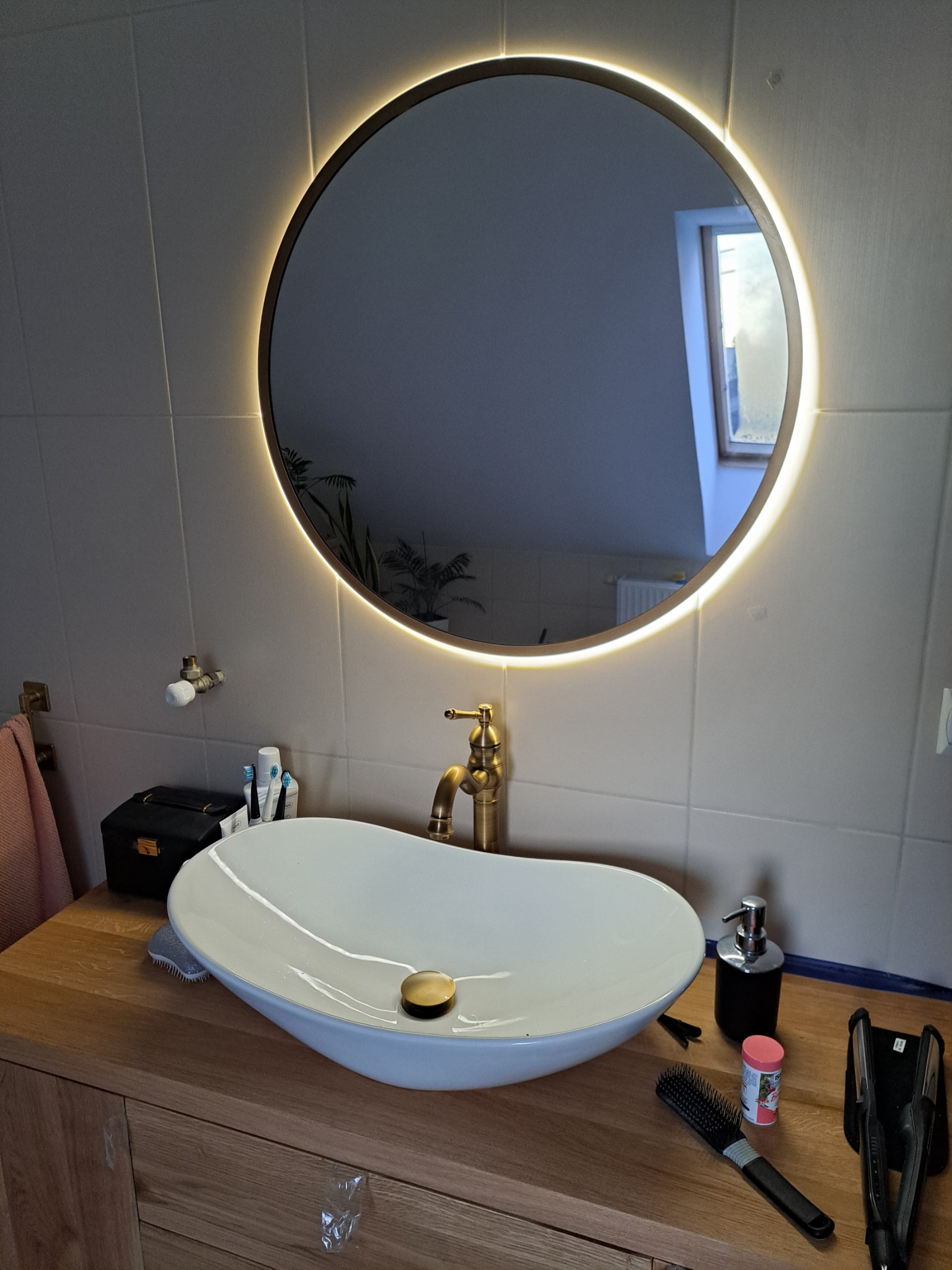 Łazienka z owalną umywalką nablatową, złotą baterią i okrągłym lustrem z podświetleniem LED, odbijającym okno dachowe.