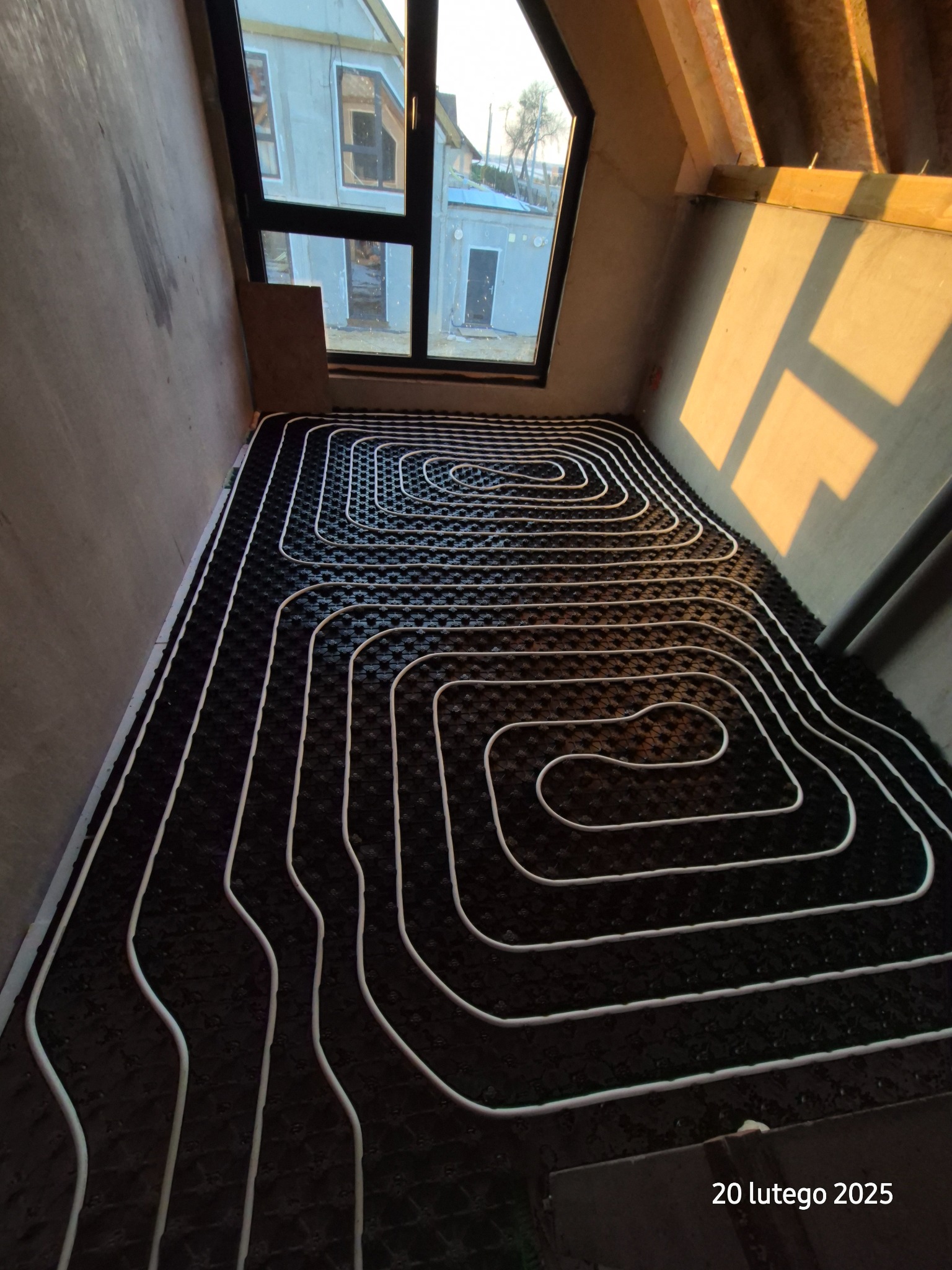 Białe rury ogrzewania podłogowego ułożone spiralnie na czarnej macie izolacyjnej w surowym pomieszczeniu z oknem, oświetlone popołudniowym słońcem.