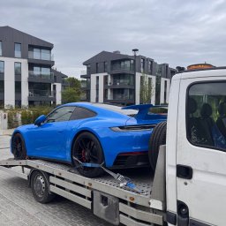 Mateusz Fras AUTOLAWETA FraHol - Błękitne Porsche na lawecie, zabezpieczone pasami transportowymi. W tle nowoczesne budynki mieszkalne i pochmurne niebo. Widoczny detal naklejki na szybie lawety.