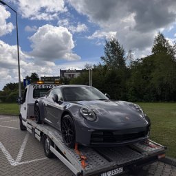 Mateusz Fras AUTOLAWETA FraHol - Szare Porsche 911 na lawecie z włączonymi światłami ostrzegawczymi, na parkingu z kostki brukowej w słoneczny dzień z chmurami.