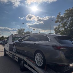 Mateusz Fras AUTOLAWETA FraHol - Szary samochód kombi na lawecie w słoneczny dzień. Widoczne tablice rejestracyjne lawety, fragment nieba z chmurami i latarnie uliczne.