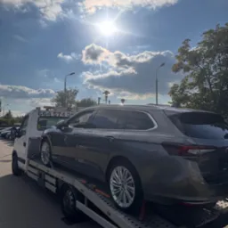Szary samochód kombi na lawecie w słoneczny dzień. Widoczne tablice rejestracyjne lawety, fragment nieba z chmurami i latarnie uliczne.