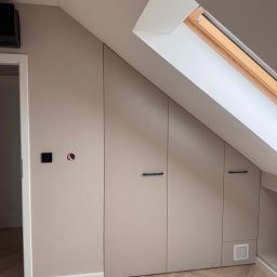 MatWood Design - Beżowa szafa wnękowa pod skosem dachu z czarnymi uchwytami, widok na parkiet w jodełkę i okno dachowe. Minimalistyczny design.
