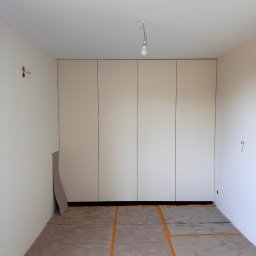 MatWood Design - Jasna szafa wnękowa z czterema drzwiami, zamontowana w pustym pokoju z przygotowanym podłożem pod podłogę. Widoczne kable i surowa żarówka.