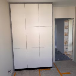 MatWood Design - Biała szafa wnękowa z szarym obramowaniem, widoczna w surowym wnętrzu z przygotowaną podłogą i otwartym przejściem do innego pomieszczenia.