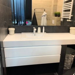 MatWood Design - Nowoczesna łazienka z białą szafką pod umywalkę z trzema szufladami, koszem na pranie i grzejnikiem drabinkowym w tle. Minimalistyczny design.