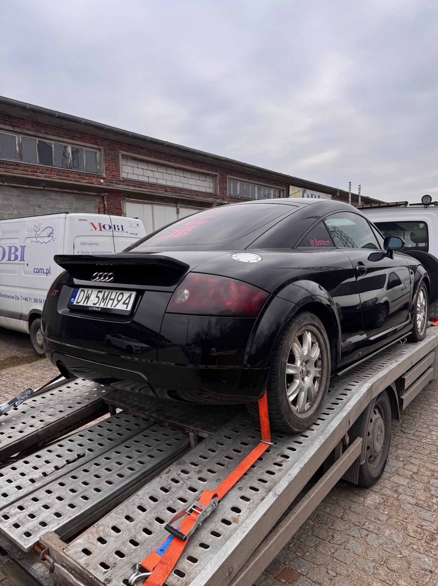 Czarny samochód Audi TT zabezpieczony pomarańczowymi pasami na platformie lawety, widoczny numer rejestracyjny DW 5MH94, w tle budynek z czerwonej cegły i biały bus z reklamą.