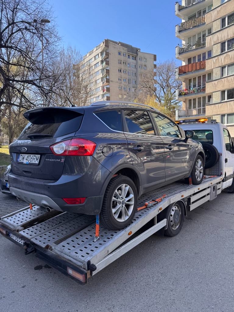 Szary Ford Kuga Titanium S, zabezpieczony pasami transportowymi, na platformie białej lawety z włączonym pomarańczowym światłem ostrzegawczym, na tle miejskiej zabudowy.