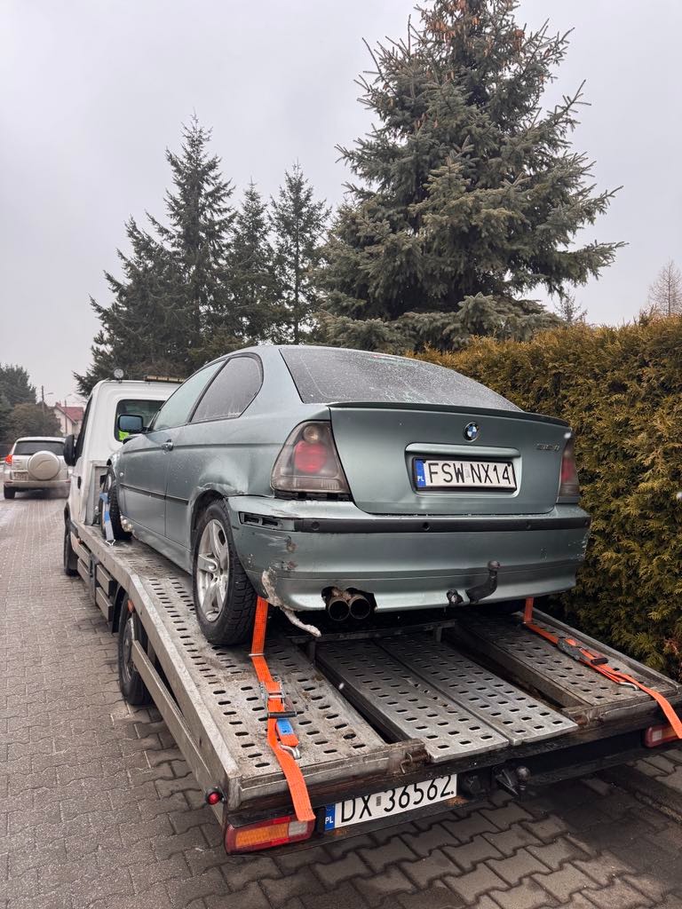 Uszkodzony, zielony samochód BMW serii 3 Coupe (E46) z polską tablicą rejestracyjną FSW NX14, zabezpieczony pomarańczowymi pasami transportowymi, przewożony na platformie lawety na tle zielonych...