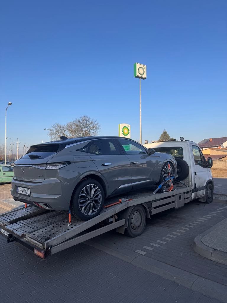 Szary samochód osobowy marki DS zabezpieczony pasami transportowymi na platformie lawety, na tle stacji benzynowej BP w słoneczny dzień.