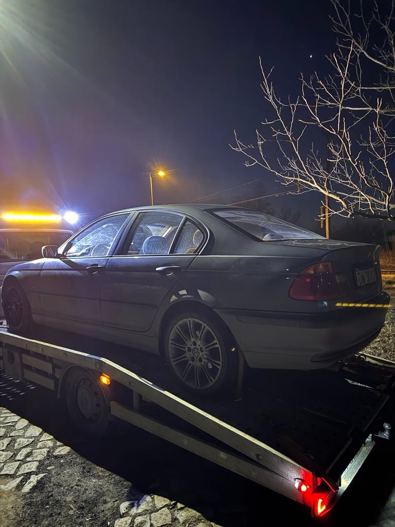 Szary samochód BMW E46 na platformie lawety, widok z tyłu, nocne ujęcie z widoczną lampą uliczną i nagimi gałęziami drzew na tle nieba.