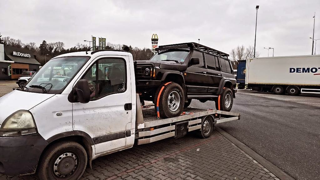 Czarny samochód terenowy z orurowaniem na dachu, zabezpieczony pomarańczowymi pasami transportowymi, przewożony na platformie lawety na tle parkingu z restauracją McDonald's.