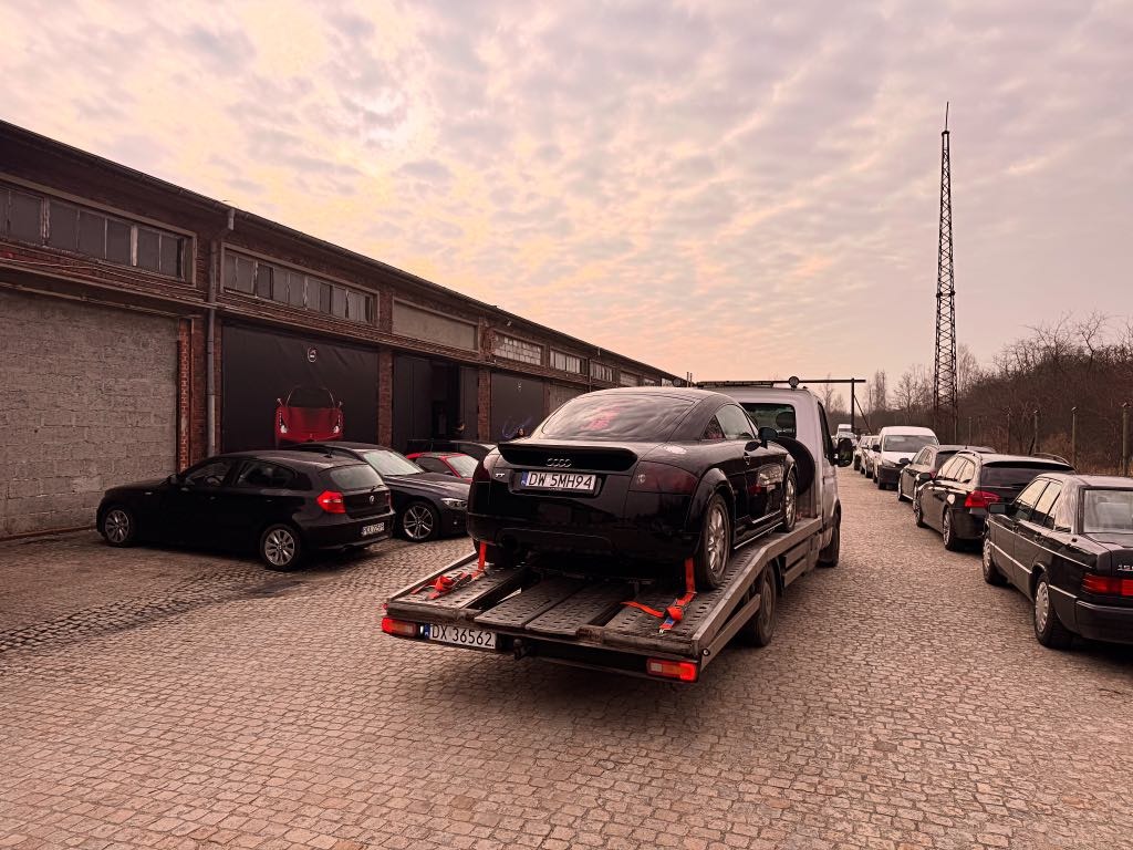 Czarny Audi TT zabezpieczony pasami na lawecie na tle ceglanego budynku z plakatem samochodu, kolejka aut oczekujących na transport.