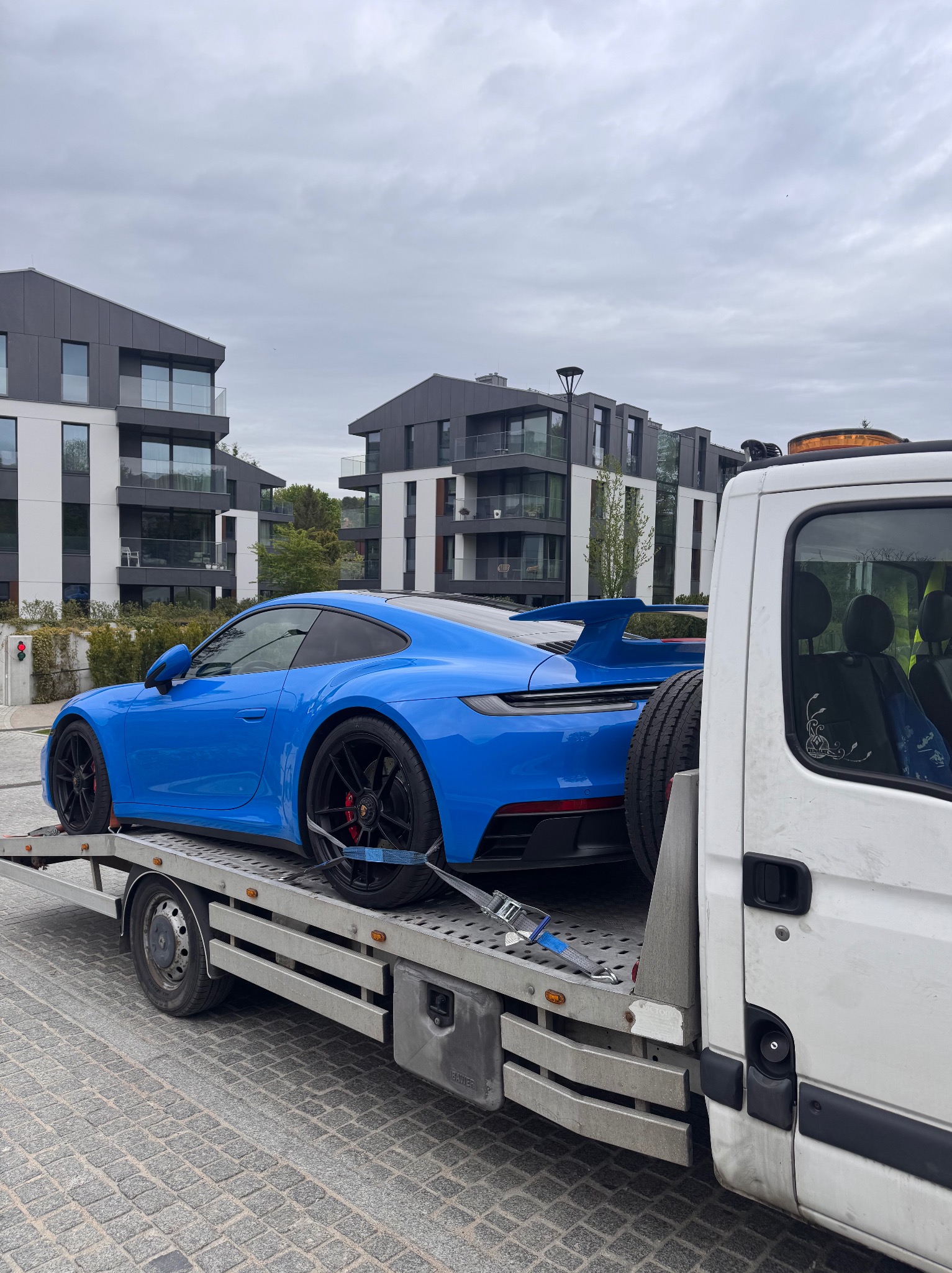 Błękitne Porsche na lawecie, zabezpieczone pasami transportowymi. W tle nowoczesne budynki mieszkalne i pochmurne niebo. Widoczny detal naklejki na szybie lawety.