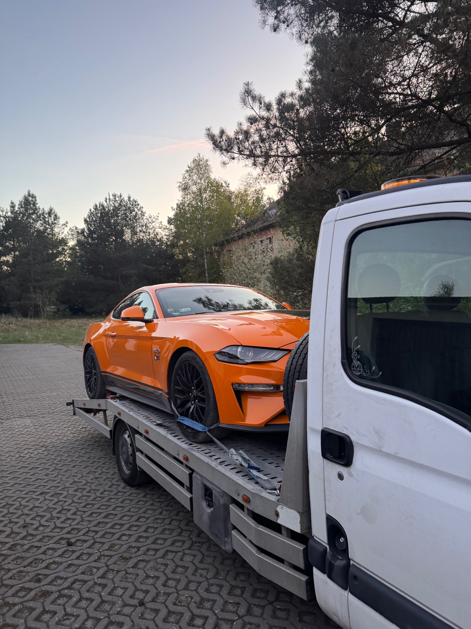 Pomarańczowy Ford Mustang zabezpieczony na lawecie w trakcie transportu, widok z boku na tle drzew i budynku, światło wczesnym rankiem.
