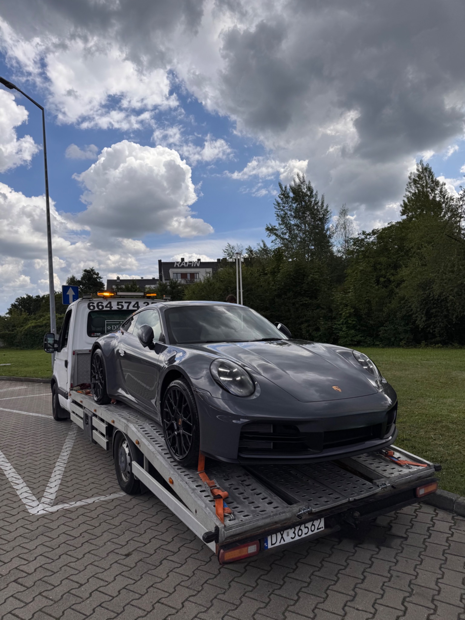 Szare Porsche 911 na lawecie z włączonymi światłami ostrzegawczymi, na parkingu z kostki brukowej w słoneczny dzień z chmurami.
