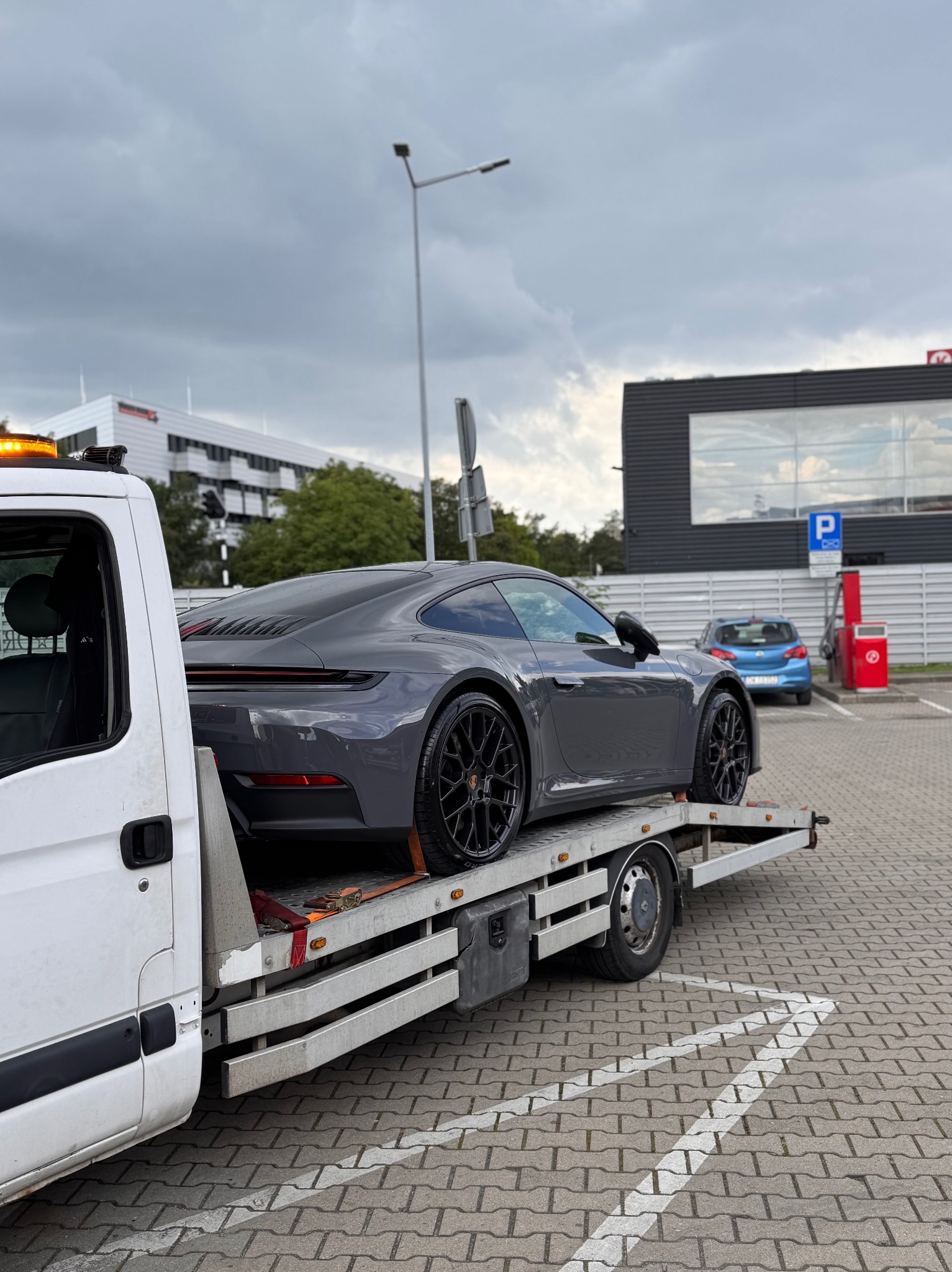 Szary Porsche 911 na białej lawecie, widok z tyłu, czarne felgi, na parkingu z kostki, budynki w tle, pochmurne niebo.