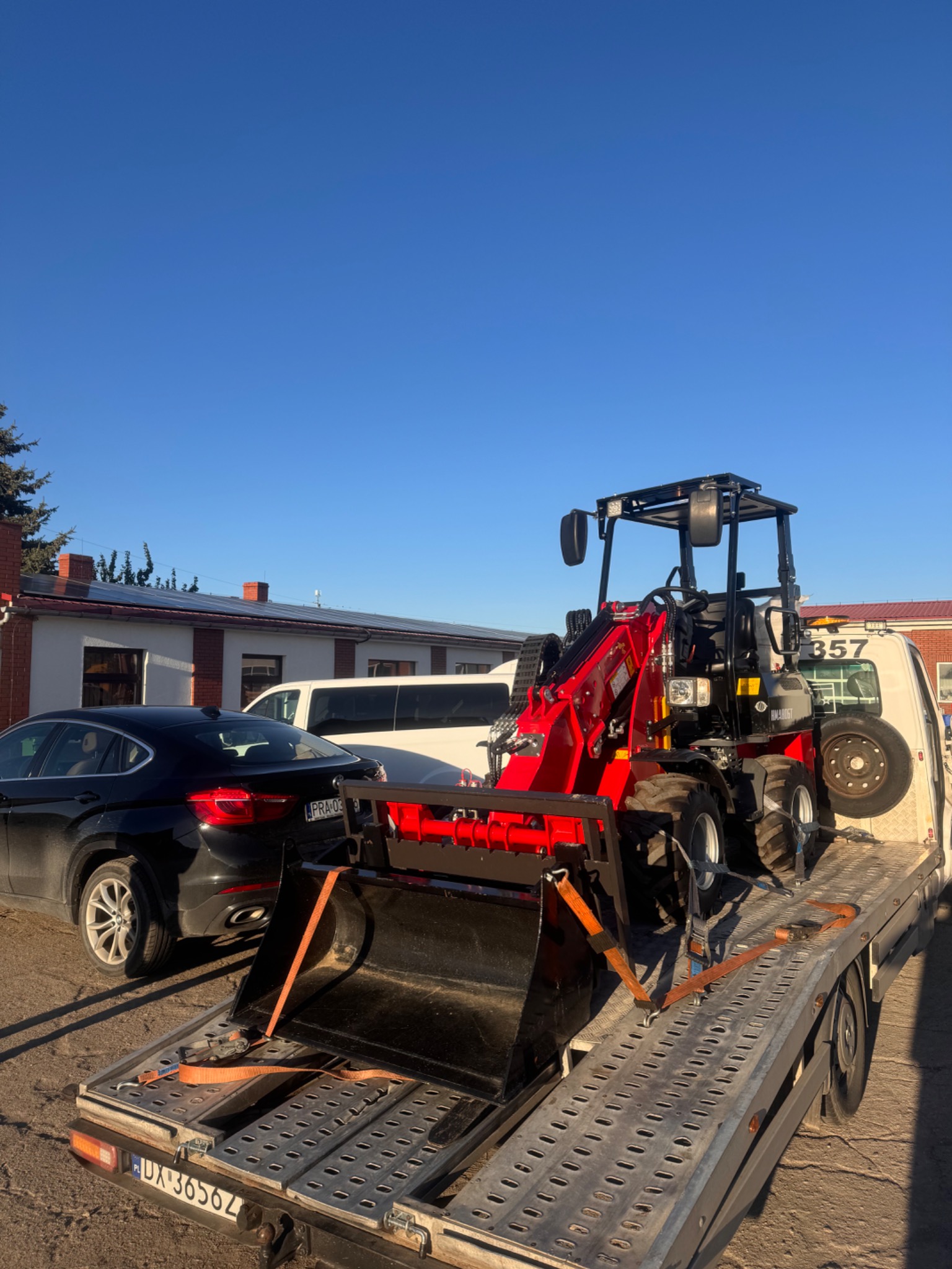Czerwona mini koparka na lawecie, zabezpieczona pasami transportowymi. W tle budynek z panelami słonecznymi i zaparkowany czarny samochód osobowy.