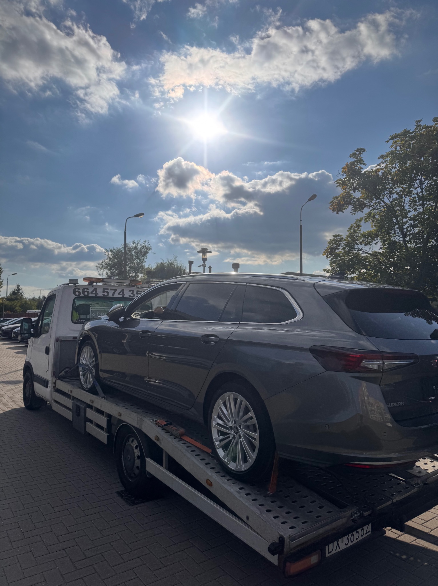 Szary samochód kombi na lawecie w słoneczny dzień. Widoczne tablice rejestracyjne lawety, fragment nieba z chmurami i latarnie uliczne.