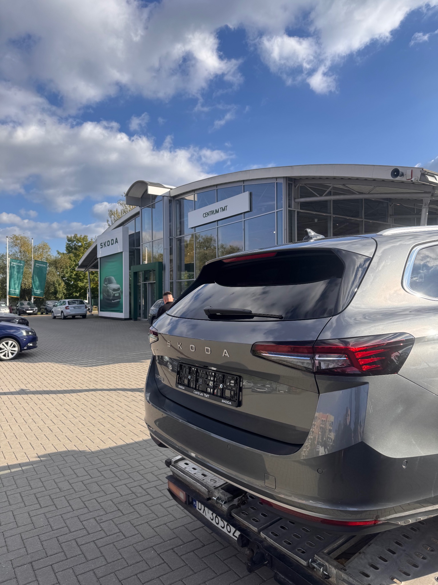 Szara Skoda Superb na lawecie przed salonem samochodowym we Wrocławiu. Widok z tyłu, błękitne niebo z chmurami w tle. Centrum TMT.