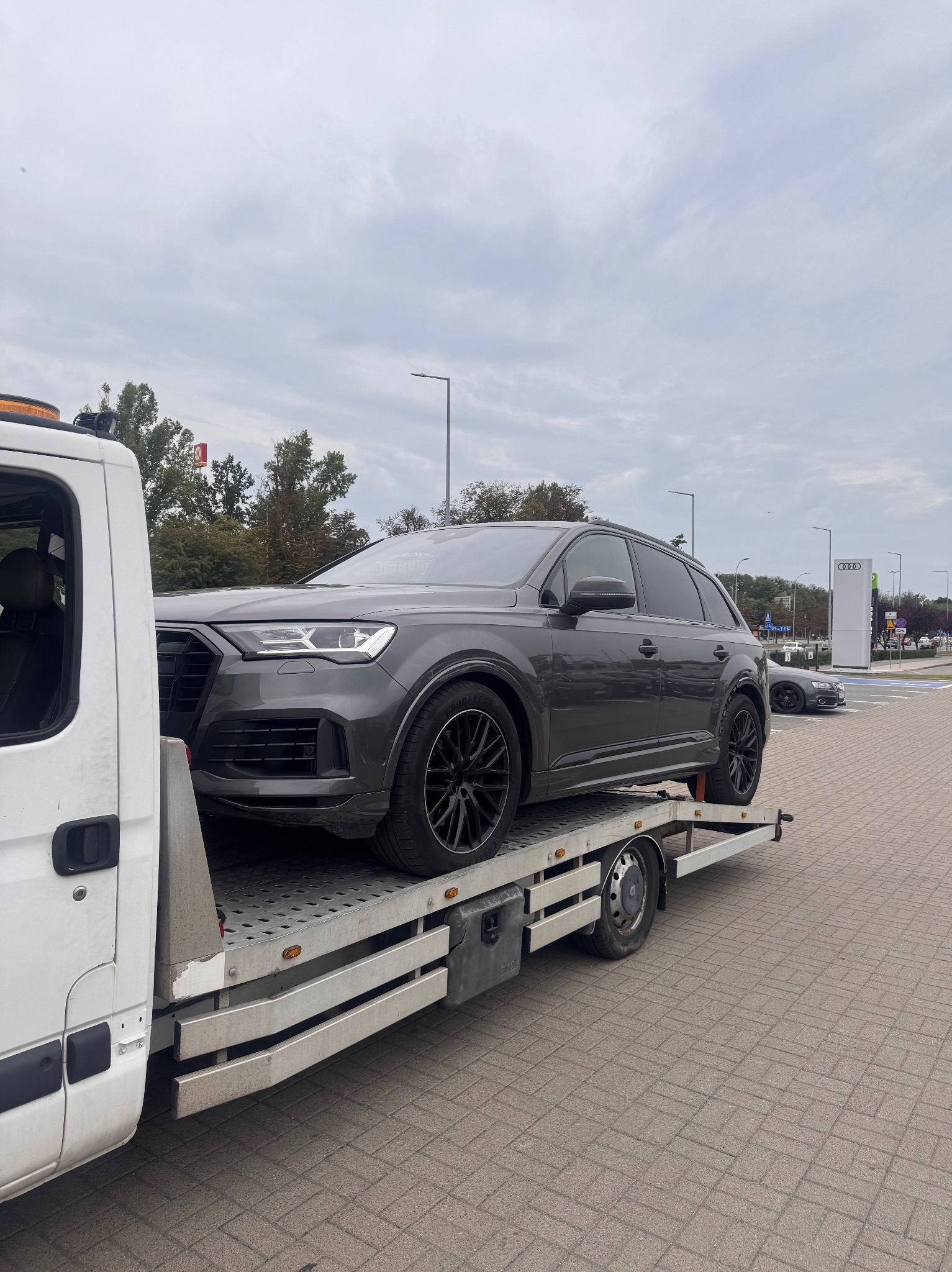 Szary SUV Audi Q7 na platformie lawety, czarne felgi, widoczny salon Audi w tle. Transport uszkodzonego pojazdu, pomoc drogowa.