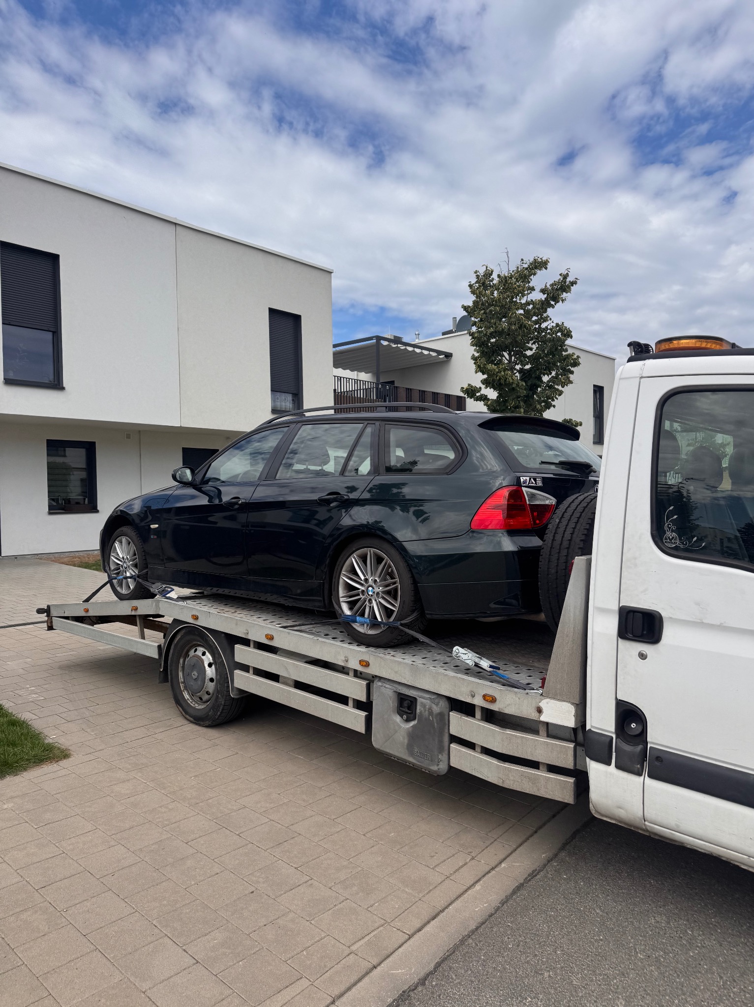 Ciemny samochód kombi marki BMW na niskopodwoziowej lawecie, zaparkowanej na chodniku z kostki brukowej, na tle nowoczesnego budynku z białymi ścianami.