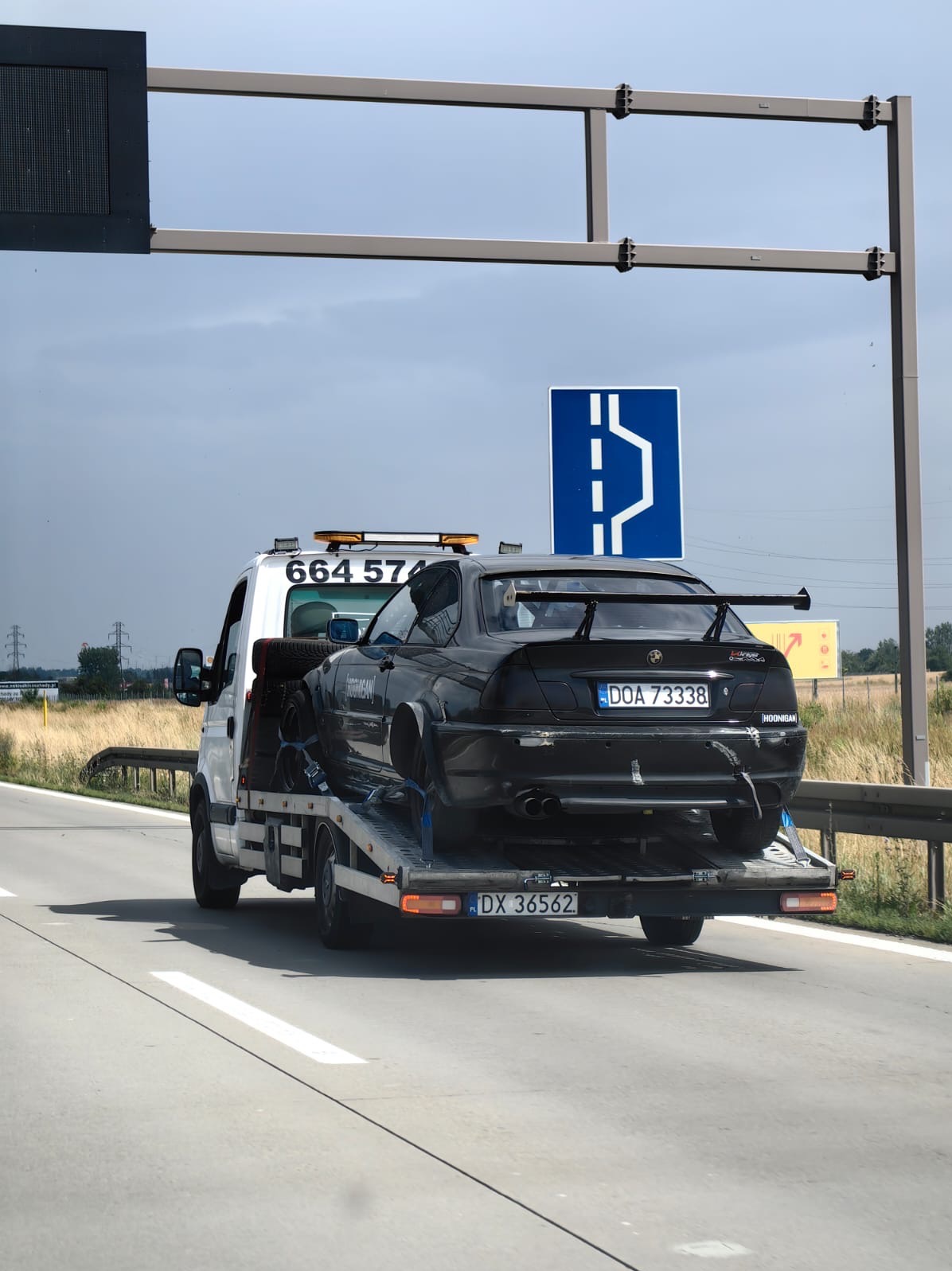 Czarna, uszkodzona sportowa BMW na lawecie, widok z tyłu. Tablice rejestracyjne DOA 73338. Laweta na autostradzie, znak drogowy nad pojazdem.