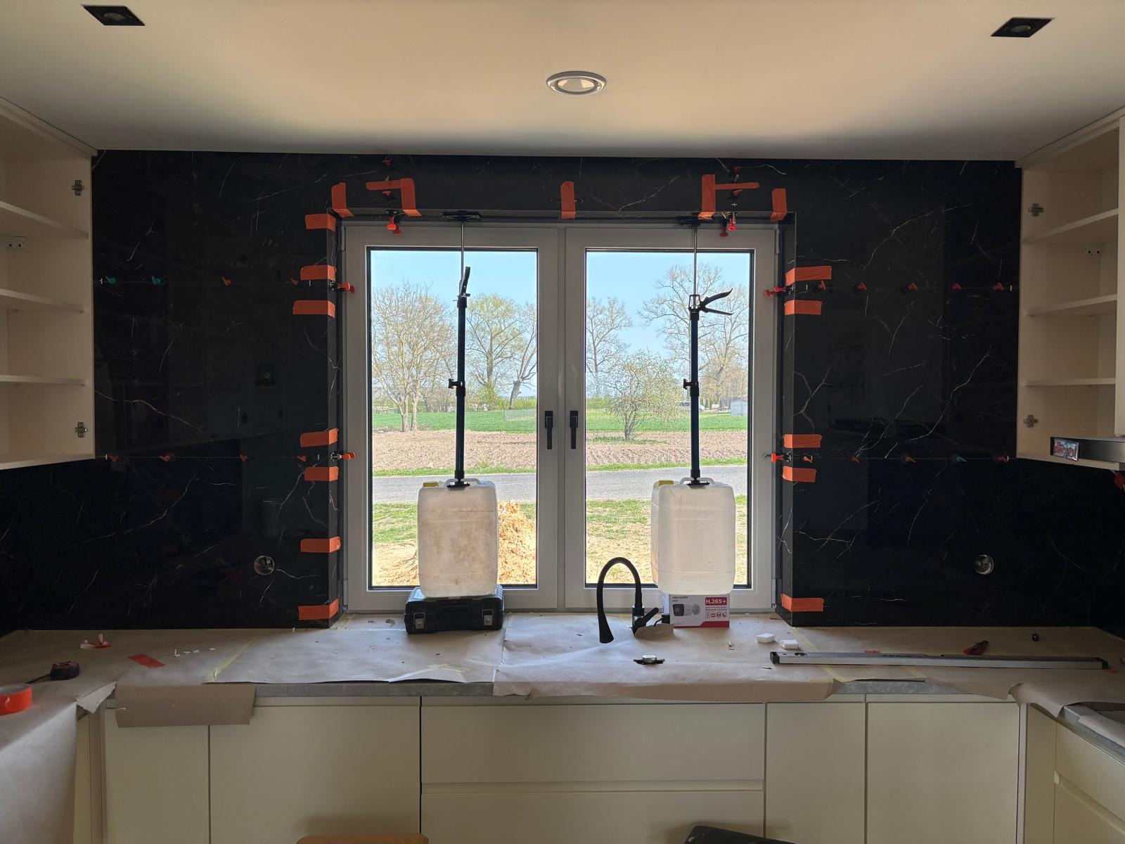 Montaż czarnych płytek z białym wzorem w kuchni, widoczne pomarańczowe klipsy poziomujące, okno z widokiem na zewnątrz i białe szafki kuchenne.