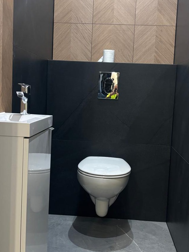 Nowoczesna toaleta podwieszana z czarną ścianą i drewnopodobnymi płytkami w górnej części, uzupełniona o umywalkę z szafką i chromowaną armaturą.