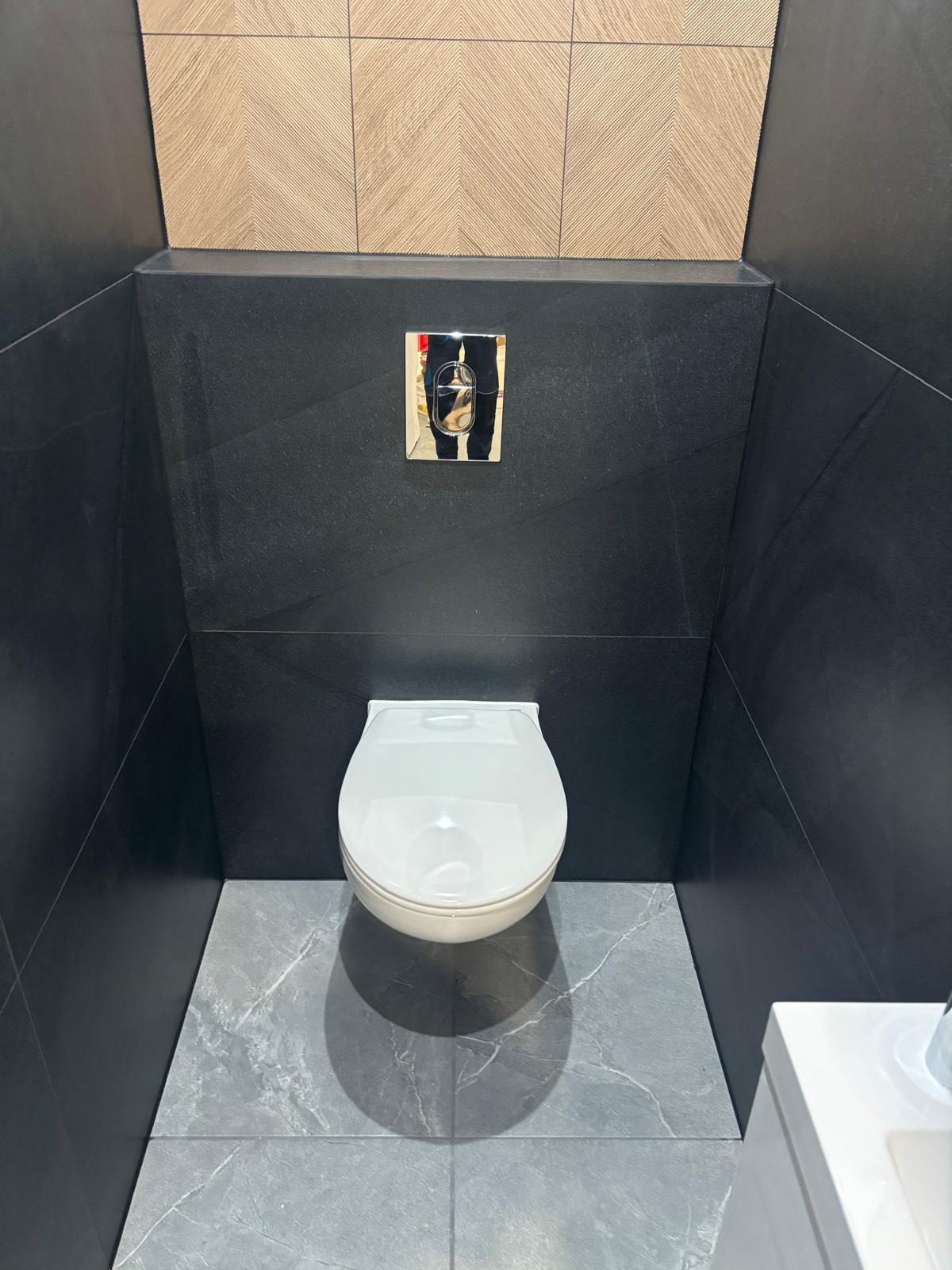 Nowoczesna toaleta podwieszana z czarnymi płytkami ściennymi i szarą marmurową podłogą. Chromowany przycisk spłukiwania. Minimalistyczny design.