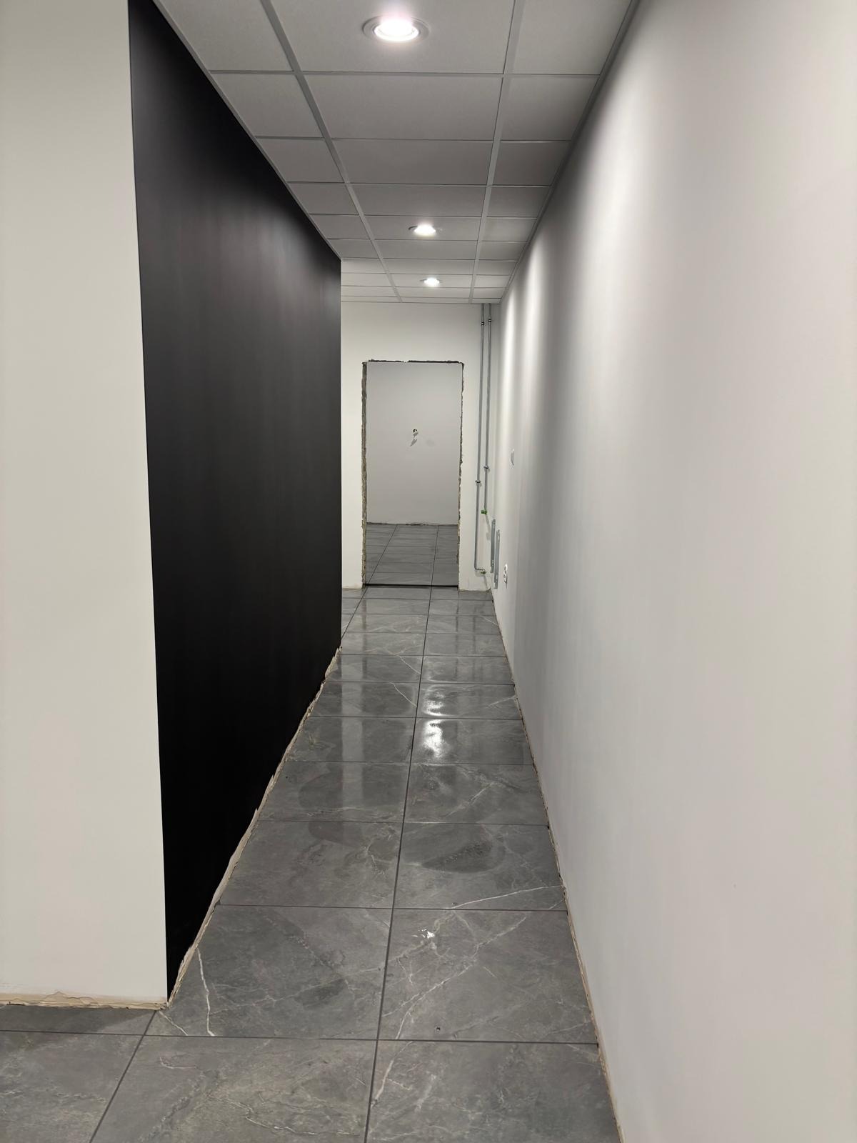 Wykończony korytarz biurowy z szarą marmurową podłogą, białą ścianą i czarną ścianą akcentującą. Widoczne oświetlenie sufitowe i otwarte drzwi w tle.