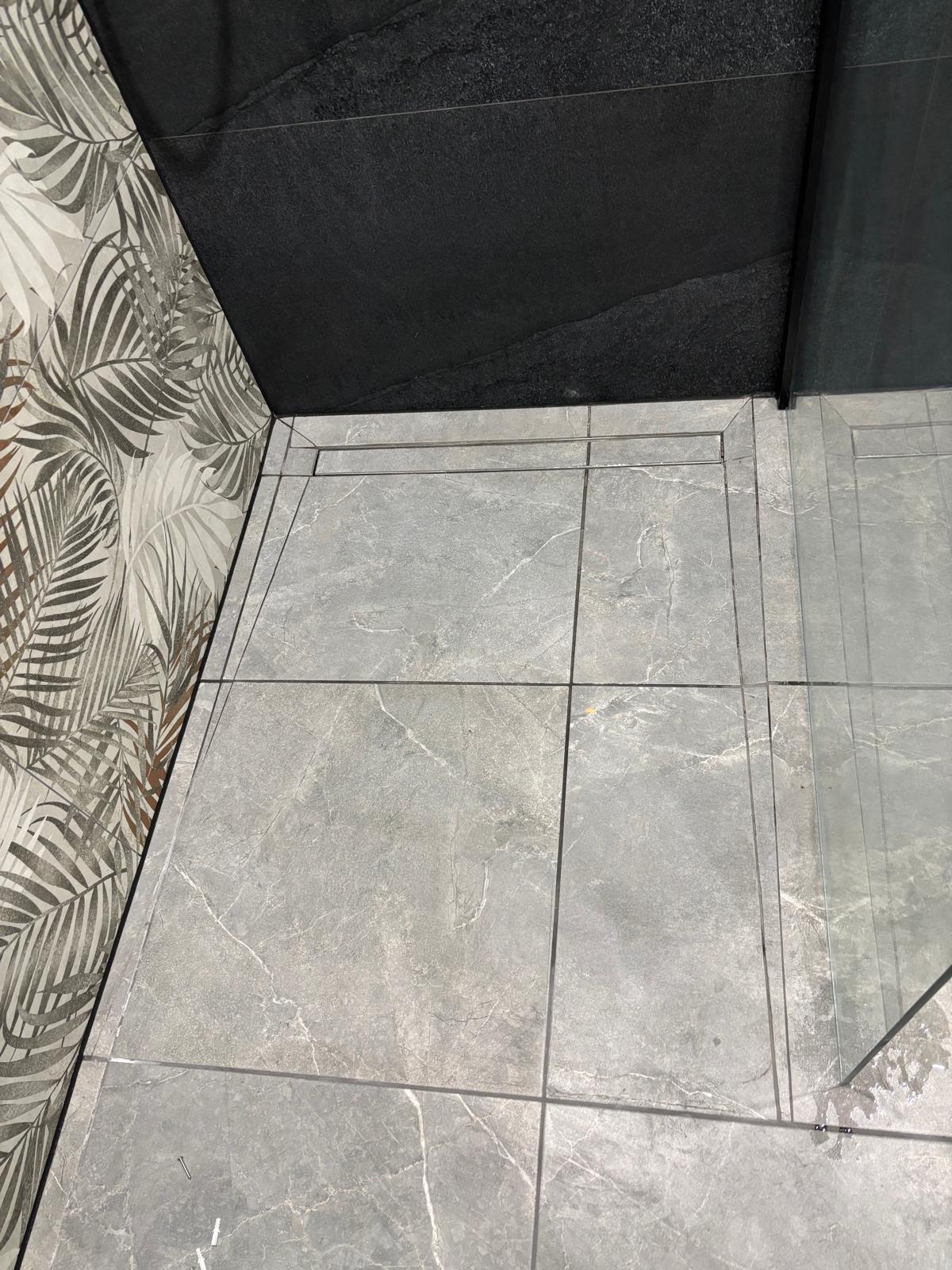 Szare płytki imitujące marmur w kabinie prysznicowej z odpływem liniowym, kontrastujące z czarną ścianą i tapetą w liście palmowe, oddzielone szkłem.