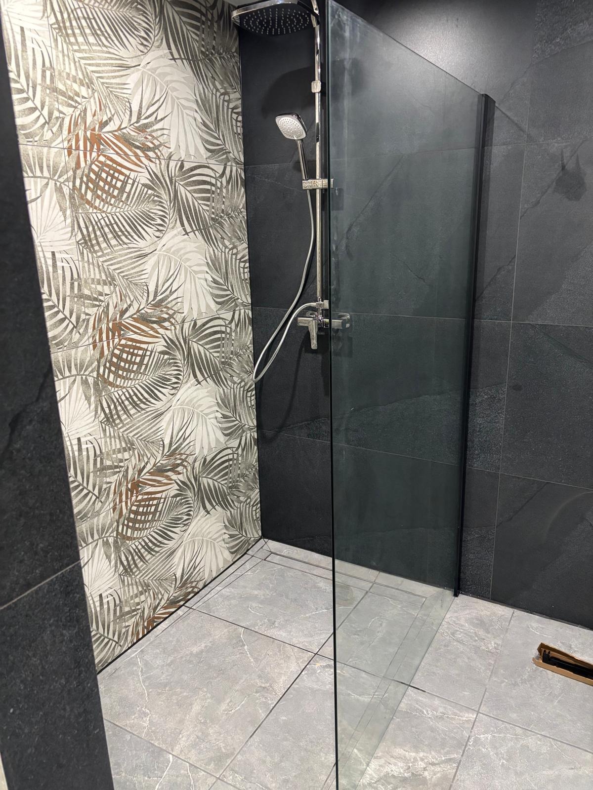 Nowoczesna kabina prysznicowa z deszczownicą, wykończona płytkami w odcieniach szarości i wzorem liści. Minimalistyczny design, szklana ścianka.